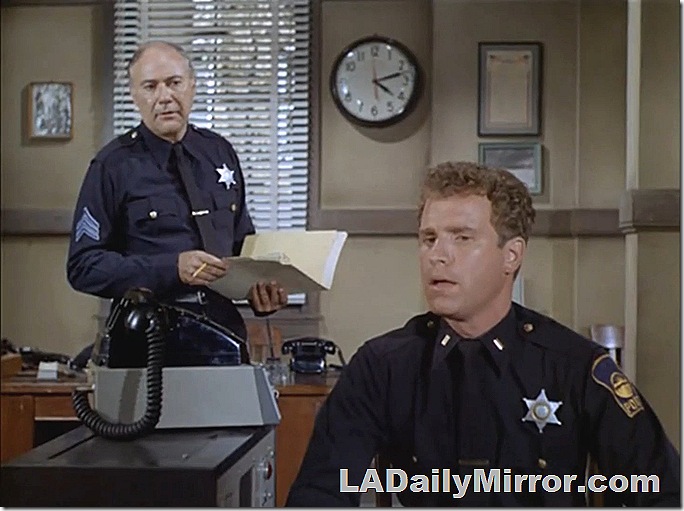 Police officer standing, holding a file. Superior officer sitting at a desk.