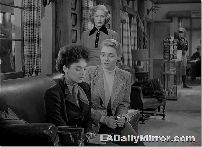 Three women. Two are sitting on a couch while a third listens in the background. 