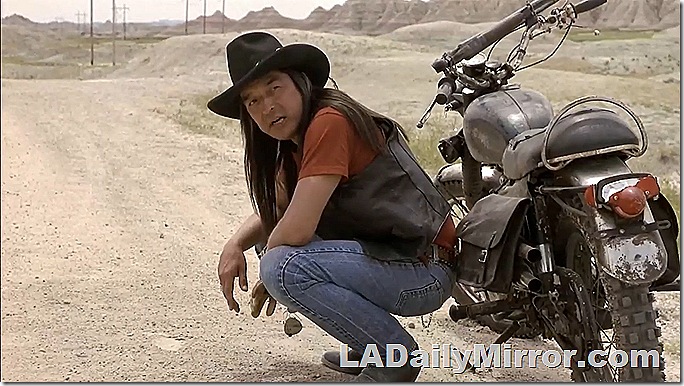 Man in jeans, vest and cowboy hat with long hair next to a motorcycle. 