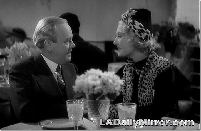 Couple sitting at table. She's wearing a coat and tie. She has a leopard print scarf and headband. 
