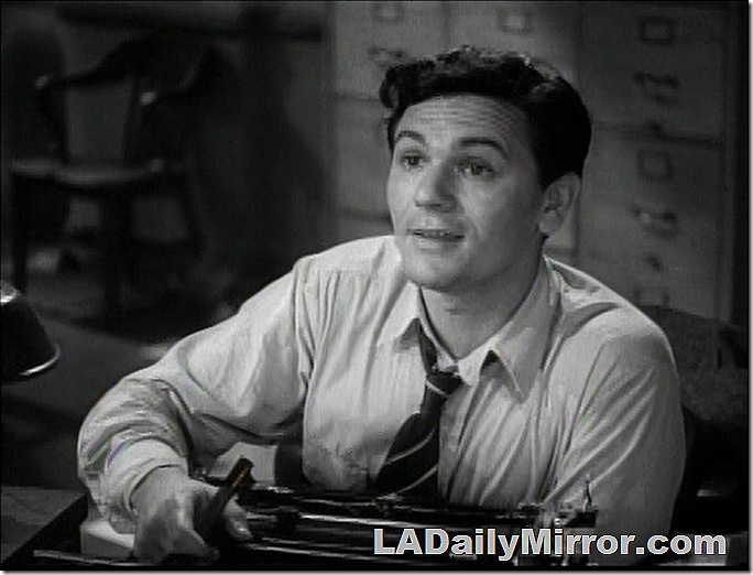 Man in shirtsleeves sitting at a typewriter. His collar is unbuttoned and his tie is pulled loose. 