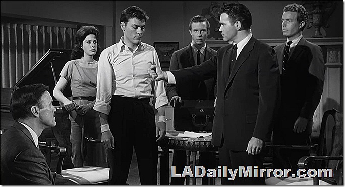 Man in a white shirt with his sleeves rolled up stands in the middle of a group of people. His wrists are bandaged. 