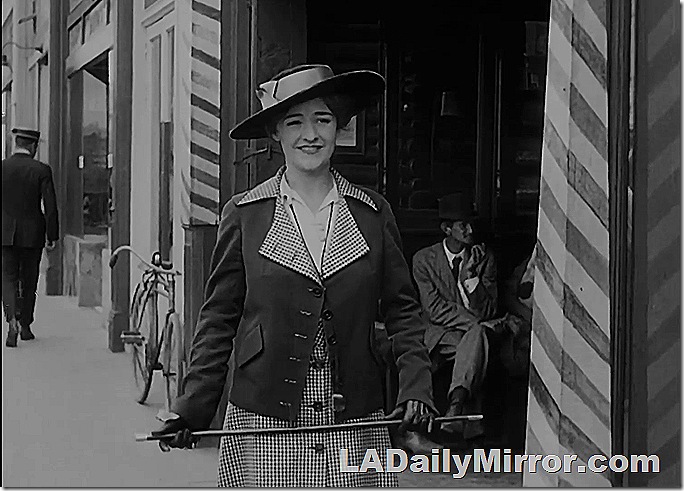 Woman in dress and coat with matching lining, and broad hat. She's carrying a cane. 