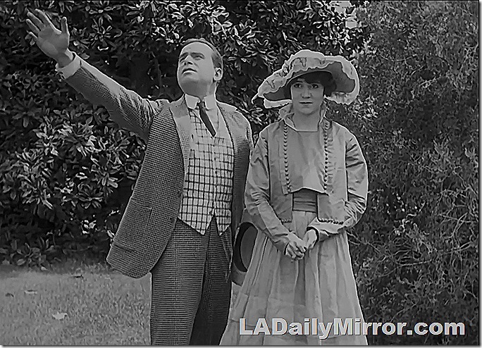 Man and woman in park. He wears a high collar and loud vest. She wears a long dress and broad-brimmed hat. 