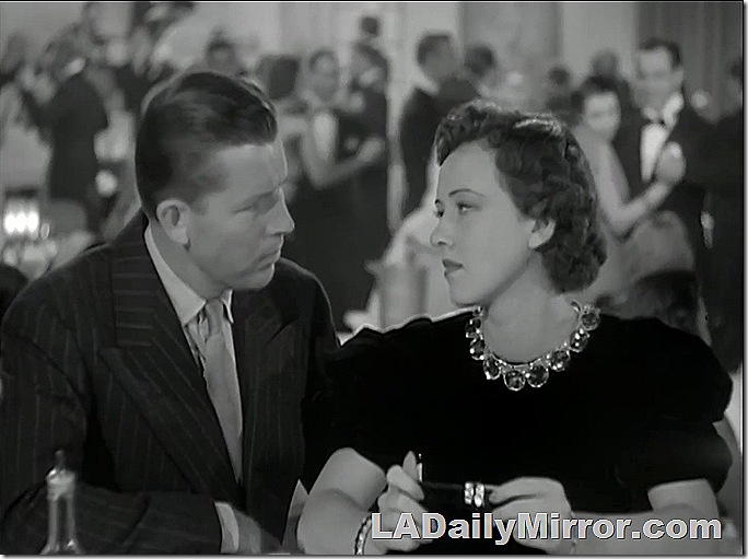 Man in double-breasted coat and tie sitting at a table with woman in elegant dress with necklace. 