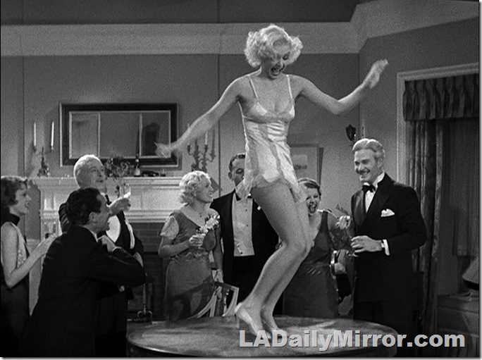 Blond woman in lingerie dancing on a table as elegantly dressed men and women watch. 