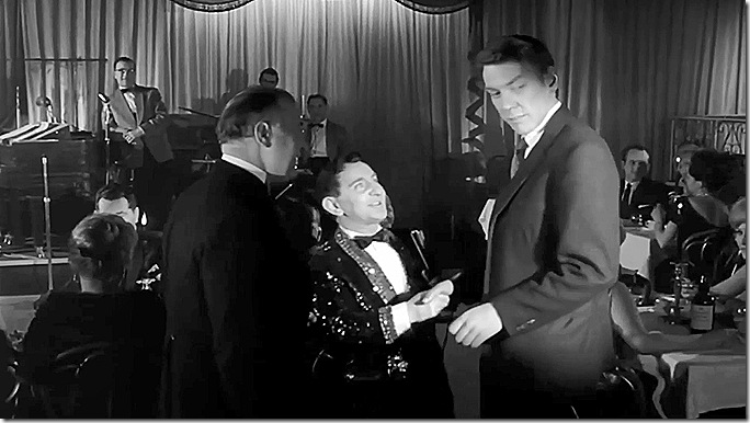 Man standing up in nightclub, talking to the emcee. 