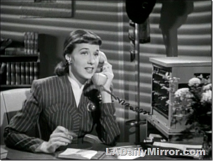 Woman with hair pulled back, in striped suit, sitting at desk and talking on the phone. 