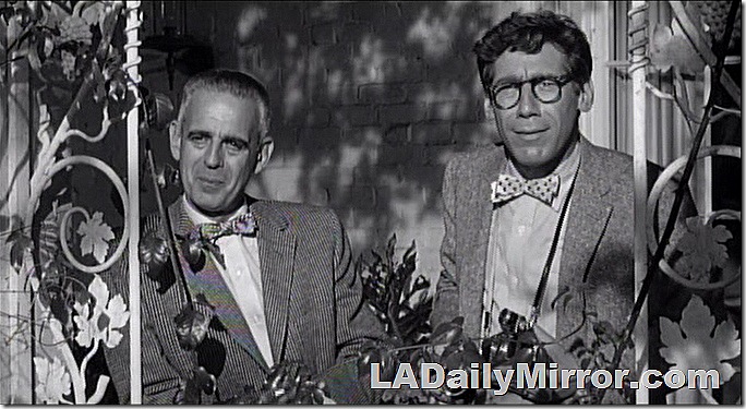 Man in coats and bow ties peering through a garden trellis. 