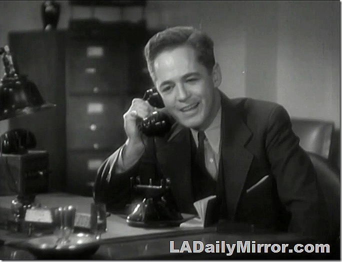 Man in coat, vest and tie, sitting at desk and speaking on a phone. 