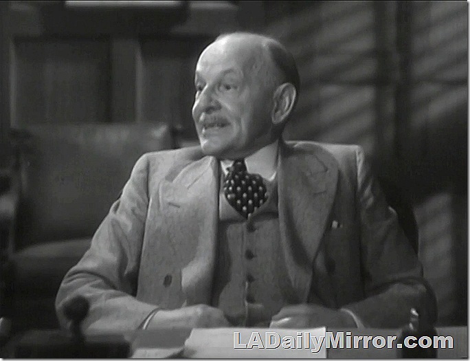 Balding man with mustache in three-piece suit sitting at desk. 