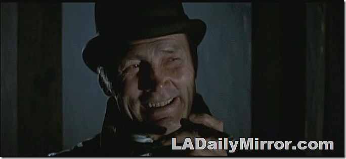 Man wearing a bowler hat, standing in shadows smoking a cigar. 