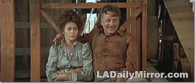 Man and woman sitting in a shed. The man has his arm around the woman. She wears and old-fashioned dress and her hair is up. He wears a shirt and suspenders. 