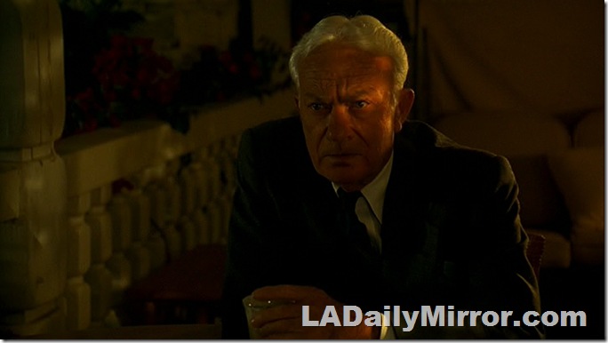 Man with white hair in coat and tie, heavily shadowed. 