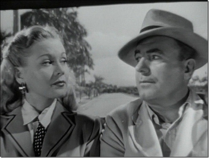 Man and woman in back seat of open car. She wears a coat, tie and stylish earrings. He was a coat and hat with an open collar.
