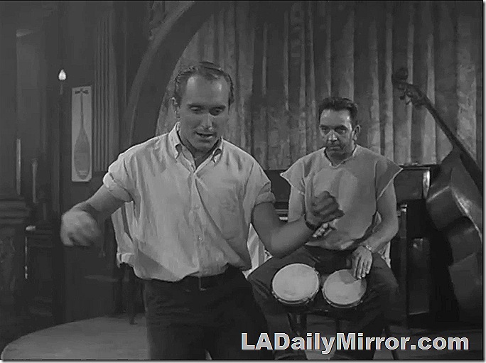 Man dances as another man plays bongos in the background. 