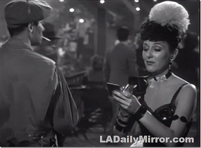 Woman in showgirl outfit with a feather on her head doing her lipstick. And Back of the Head Guy. 