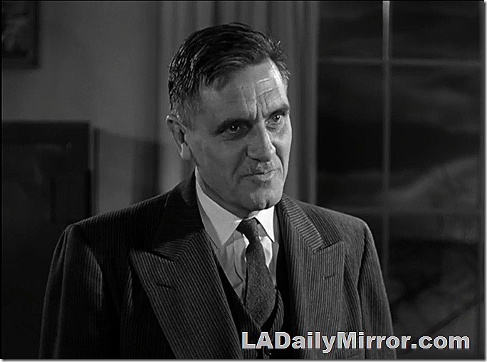 Man with mustache, in three-piece suit, standing next to window. 