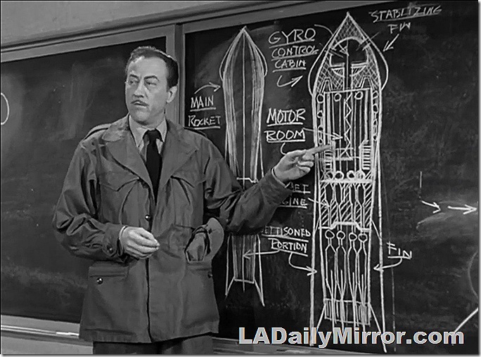 Man in Army field jacket next to elaborate drawing of a spaceship on a blackboard. 