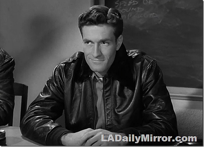 Man in leather flying jacket sitting at a table, half-smiling