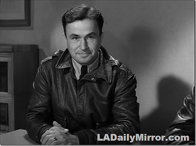 Man sitting at table, wearing leather flying jacket and tie. 