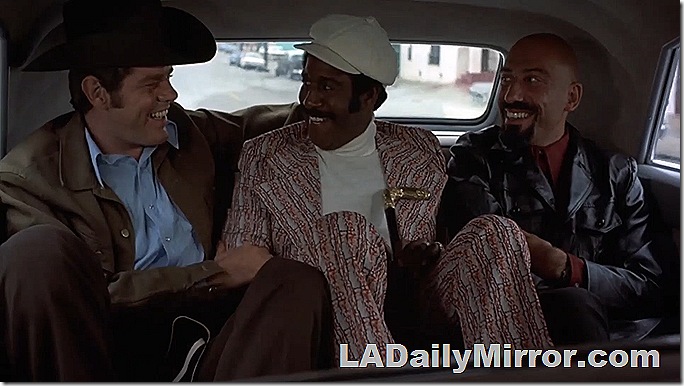 Three men in the back of a limo. One wears a cowboy hat, another is bald and the man in the middle has a suit with wide lapels and carries a cane. 
