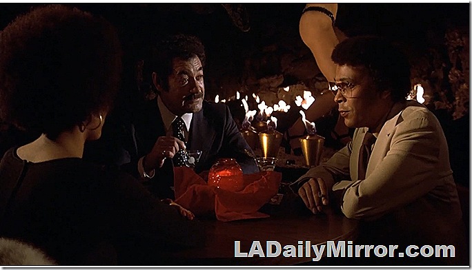 Men sitting at a table in a topless bar. A dancer is in the background. 