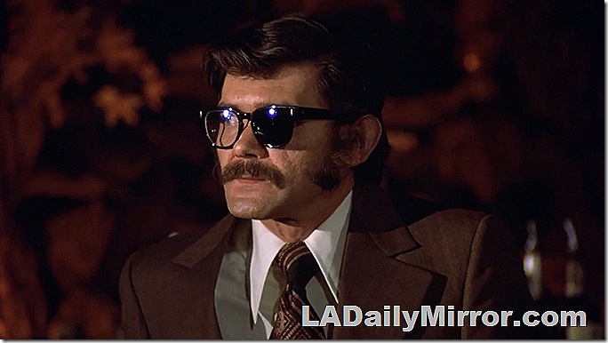 Man with mustache in suit, he wears glasses and the left lens is covered. 