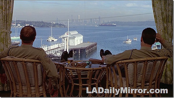 Two men sitting in chairs with their feet up, looking at a view of the ocean with a city in the distance. 