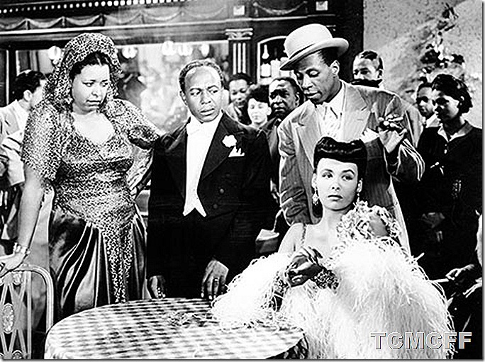 Nightclub scene with a woman in a feathery outfit sitting at a table as a woman and two men stand behind her. 