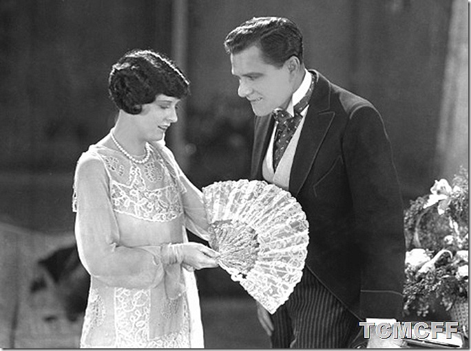 Woman holding fan as a man looks at her. 
