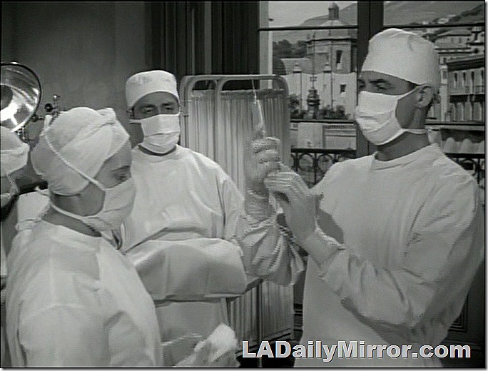 Surgical scene. A surgeon in cap, mask and gown examines a hypodermic needle as a doctor and nurse watch. 