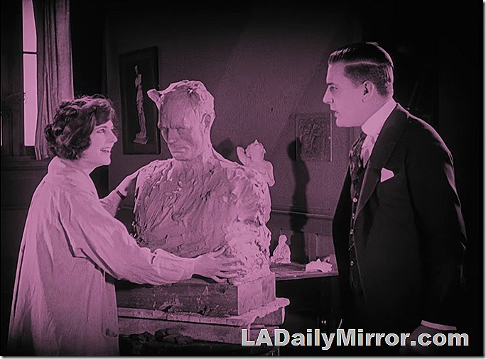 A woman in an artist's smock shows off a scowling sculpture to a man in a suit and tie. A woman in an artist's smock shows off a scowling sculpture to a man in a suit and tie.