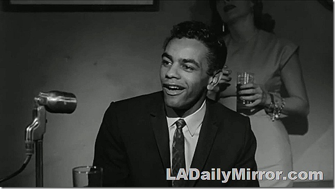 Man in coat and tie next to a microphone. A woman is standing in the background and holding a drink. Man in coat and tie next to a microphone. A woman is standing in the background and holding a drink.