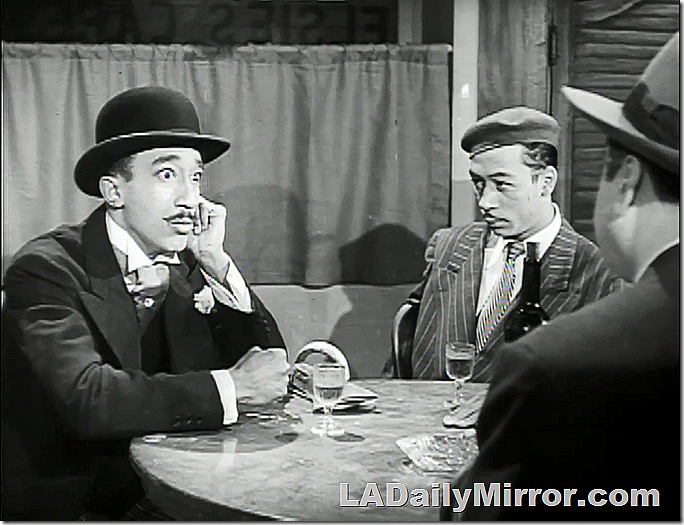 Three men sitting at a table. One has a slight mustache, wears a derby and a wing collar. The second wears a beret and a pinstripe suit. The third has his back to the camera. 