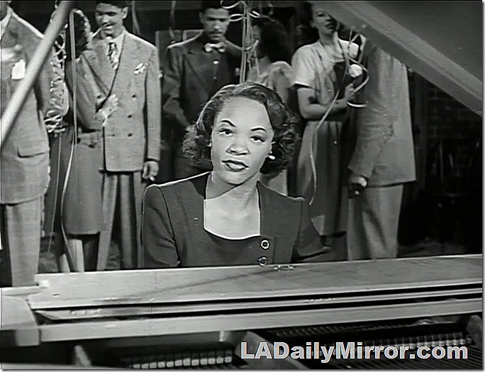 Woman in fashionable dress playing the piano, looking at the camera. 