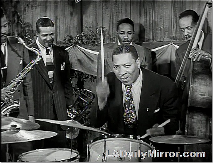 Man in coat and tie playing the drums. The rest of the band watches him play. Man in coat and tie playing the drums. The rest of the band watches him play.