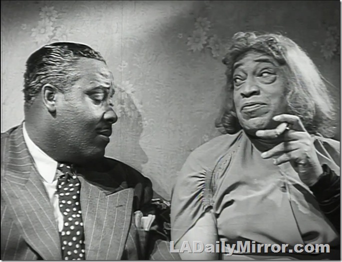 Two people sitting on a couch. The man on the right wears a pinstripe suit. The person on the left has long hair and smokes a cigar. Two people sitting on a couch. The man on the right wears a pinstripe suit. The person on the left has long hair and smokes a cigar.
