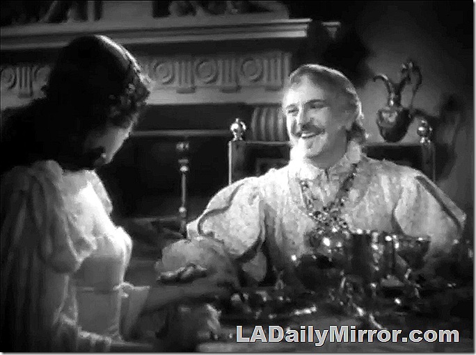 Richly dressed man with Van Dyke beard at a table with elaborate settings. He pats a woman on the arm. 