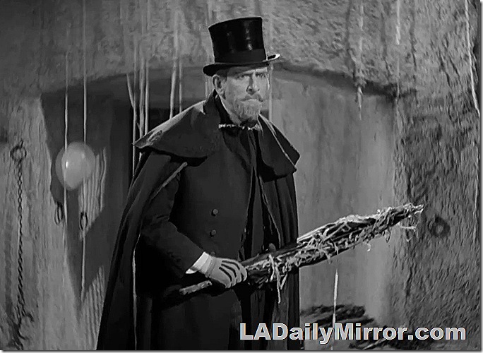 Bearded man in top hat and cape holding an umbrella in a menacing fashion. 