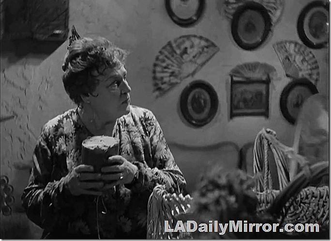 Older woman with hair pulled up, in a robe, holding a loaf of bred. The wall is decorated with fans. 