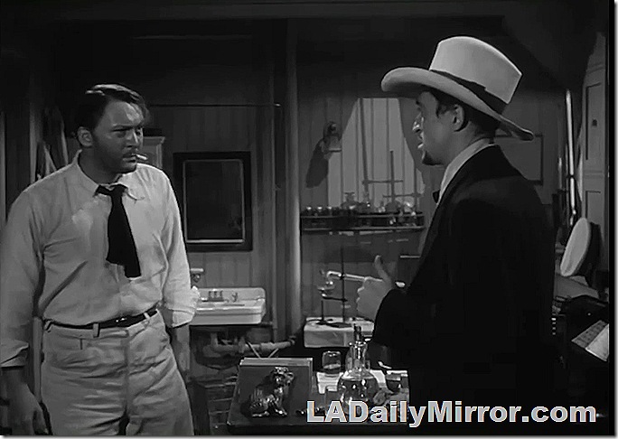 Man smoking cigarette, with short necktie, looking at man in coat and hat. 