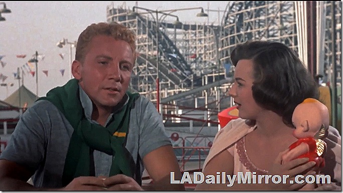 A roller coaster! With a couple in the foreground. The man has a sweater tied around his neck. The woman is holding a doll. A roller coaster! With a couple in the foreground. The man has a sweater tied around his neck. The woman is holding a doll.
