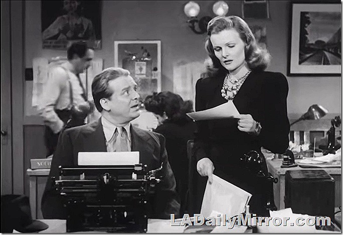 Man in an office, wearing a coat and tie sitting a typewriter, looking at woman who is holding a piece of paper. 