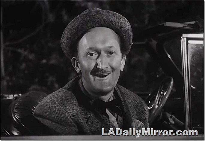 Man in coat and bow tie with slight mustache, in seat of car wearing hat with brim pushed back in comical fashion. 