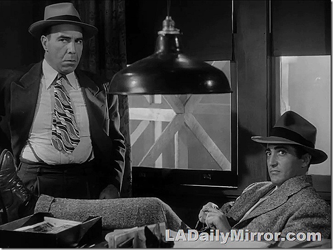 Men in coats and hats. One is sitting at a desk with his feet up. The other one has his jacket pulled back. 