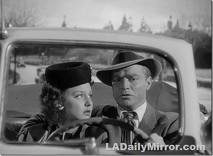 Man in suit and hat, woman in dress and hat, riding in a convertible with the top down. 