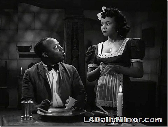 Man sitting at table in jacket shirt and bow tie, looking at woman standing nearby in maid's outfit. Man sitting at table in jacket shirt and bow tie, looking at woman standing nearby in maid's outfit.