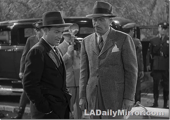 Two men in suits and hats, outdoors with a car and police officers in the background. 