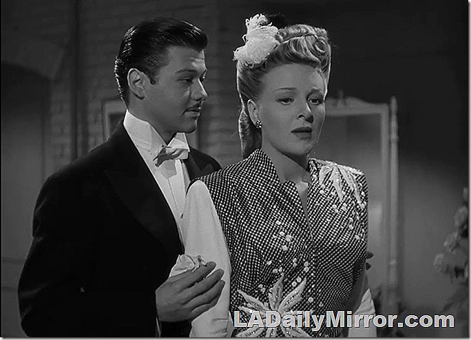 Man in tuxedo standing behind woman in dazzling outfit. 